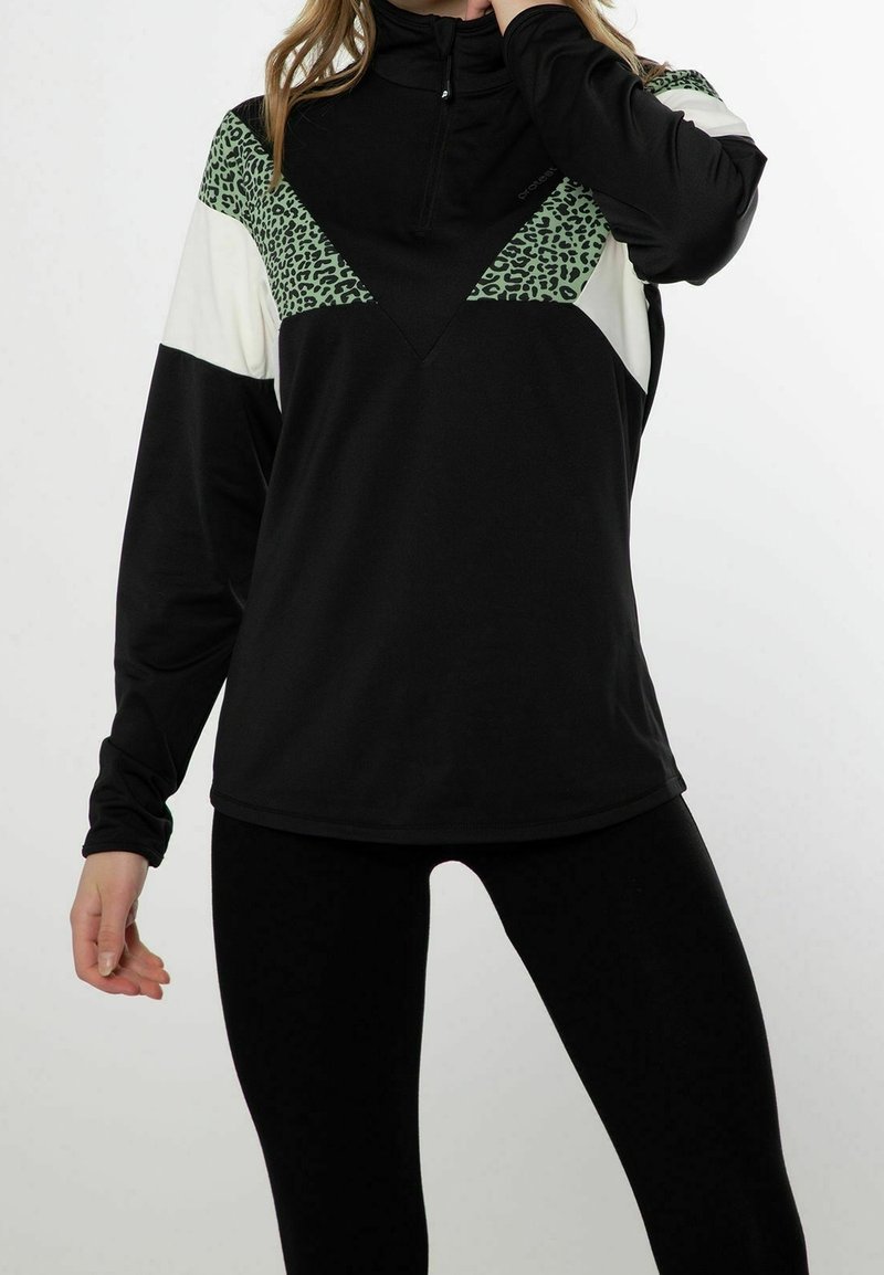 Femme portant des leggings noirs et un pull noir avec un imprimé léopard vert et des panneaux blanc cassé en blocks de couleur, posant avec une main près du cou.