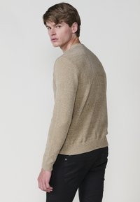 Bež pleteni pulover z strukturo, okroglo izrezana vrata, rebrasti manšeti in rob, ter prilegajoča silhueta. Model nosi črne hlače.