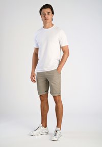 Camiseta blanca de algodón, pantalones cortos beige a cuadros con puños enrollados y zapatillas deportivas blancas con cordones multicolores. Pose de pie.
