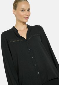 Woman with slicked-back hair wearing a black long-sleeve buttoned blouse with lace detail across the chest, smiling slightly.