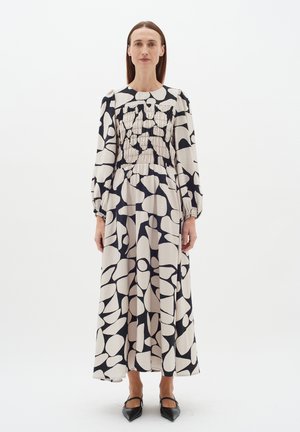 Maxime vestido em creme e preto com padrão abstrato, corpete franzido, mangas longas bufantes e faixa elástica na cintura. Usado com sapatos pretos de bico fino.