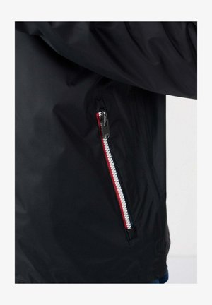 Giacca impermeabile nera con tasca laterale con cerniera a contrasto bianca e accenti rossi, tessuto strutturato e design moderno.
