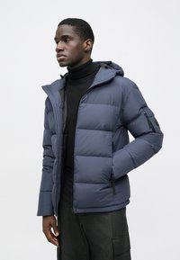 Sötétkék pufikabát kapucnival, steppelt részletekkel, cipzáras záródással és két oldalzsebbel; fekete garbó fölött viselve.