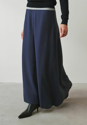 Frau trägt weite marineblaue Hosen, schwarze spitze Stiefel mit Absatz, schwarzes langärmeliges Oberteil und silbernen Ring an der rechten Hand.