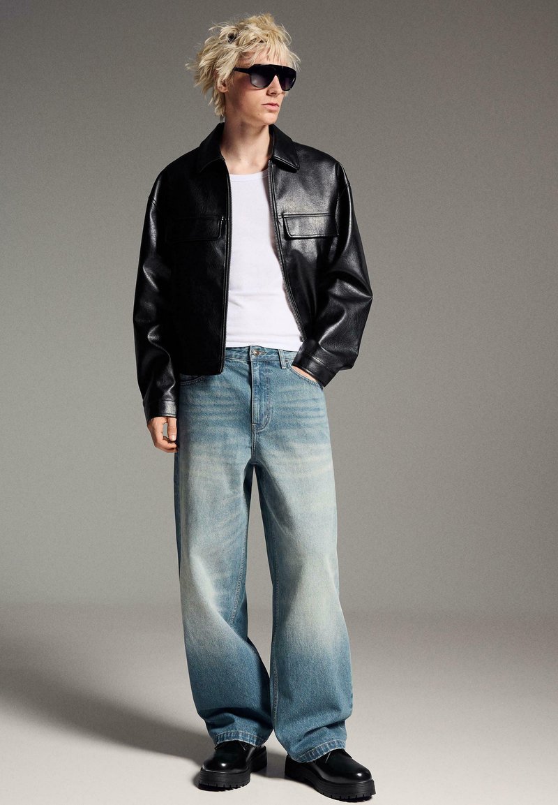 Personne blonde portant une veste en cuir noire surdimensionnée, un t-shirt blanc, un jean bleu clair ample, des chaussures noires épaisses et de grandes lunettes de soleil noires.