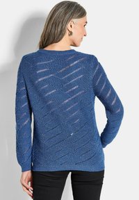 Blauer Strickpullover mit einem strukturierten, offenen Muster und langen Ärmeln. Hat einen runden Ausschnitt und einen gerippten Saum, kombiniert mit schwarzen Hosen.
