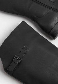 Botas de cuero negro hasta la rodilla con una textura suave, cremalleras laterales y una correa de acento con detalle de hebilla.