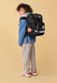 Black backpack with a lightning bolt pattern, side pockets, and colorful badges, worn on a child's back with a denim jacket and striped pants.