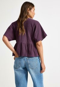 Fialový texturovaný top s krátkými rukávy a peplum zakončením, zkombinovaný se světle modrými širokými džíny. Zadní pohled ukazuje střih a styl oděvu.