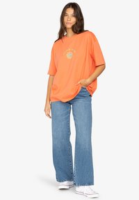Orange oversized t-shirt with short sleeves, featuring a circular graphic design in the center. Paired with blue flared jeans and white sneakers.