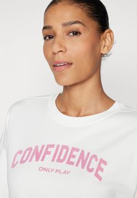 Sweatshirt blanc avec un col rond et un grand imprimé rose portant la mention "LA CONFIANCE SEULEMENT JOUER." Tissu lisse et coupe décontractée.