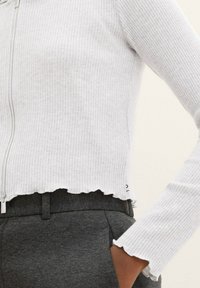 Biały prążkowany krótki top z zamkiem z przodu, z postrzępionymi brzegami i długimi rękawami, zestawiony z ciemnoszarymi spodniami. Tekstura wygląda na miękką i dopasowaną.