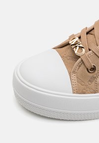 Gros plan d'une basket en daim beige avec embout en caoutchouc blanc, semelle blanche épaisse, lacets beiges et breloque logo dorée sur les lacets.