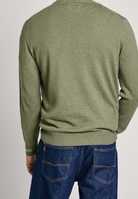 Grüner Strickpullover mit rundem Ausschnitt, langen Ärmeln und gerippten Bündchen, kombiniert mit blauen Jeans aus Denim mit orangefarbener Steppnaht.