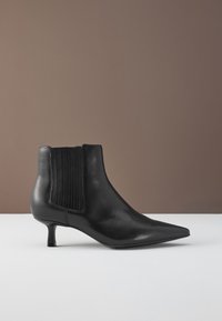 Bottine en cuir noir à bout pointu, petit talon kitten et panneau latéral élastique sur une surface claire avec un fond marron.