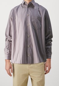 Camicetta button-up di colore viola con righe bianche verticali. Presenta una tasca sul petto, maniche lunghe e un colletto classico. Tessuto dalla superficie liscia.