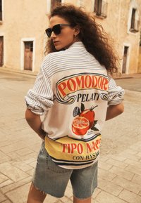 White shirt with blue stripes featuring a colorful "Pomodoro" graphic and yellow accents, paired with distressed denim shorts.