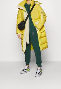 Keltainen puffer-takki valkoisilla yksityiskohdilla, vihreä collegeasu ja musta crossbody-laukku. Valkoiset ja mustat lenkkarit neonkeltaisilla yksityiskohdilla.