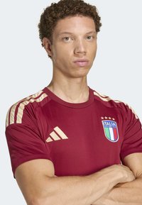 Mladić s kovrčavom kosom nosi bordo nogometnu majicu Italije s zlatnim Adidas trakama i logotipom, prekriženih ruku, neutralnog izraza lica.