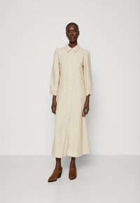 Bežs pogu krekla kleita ar apkakli, garām puff piedurknēm un vidējā garuma svārkiem. Modelis valkā brūnas papēžu mules. Gluda auduma tekstūra.