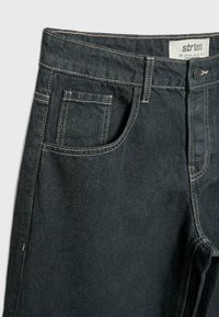 Gros plan sur un jean en denim noir avec surpiqûres blanches visibles, poche avant, poche à monnaie, passants de ceinture et étiquette de marque sur la ceinture.