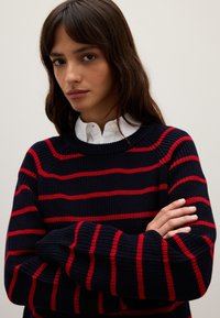 Maglione a costine blu navale con strisce orizzontali rosse, con scollo a giro e maniche lunghe, indossato sopra una camicia bianca con colletto.