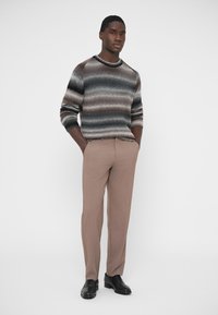 Dryžuotas megztinis žemės atspalvių, derinamas su smėlio spalvos kelnėmis. Avalynė – juodos odos. Audinys atrodo struktūruotas ir šiltas.