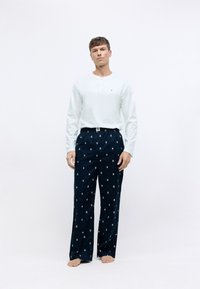 Chemise henley blanche à manches longues assortie à un pantalon noir à motifs avec des accents répétés en bleu clair et rose, présentée dans un cadre minimal.