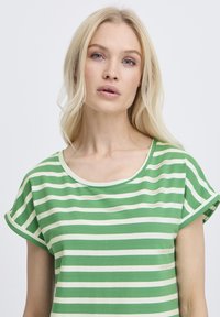 Szőke nő világoskék szemekkel, zöld-fehér vízszintes csíkos rövid ujjú felsőt visel, elölről néz a sima háttér előtt.