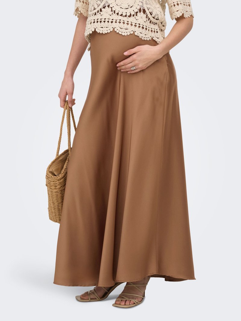 Femme portant une jupe longue marron et un top en crochet, tenant un sac à main tissé, avec des sandales à lanières, la main reposant sur son ventre.