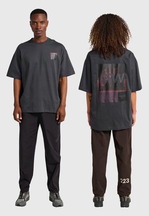 Två modeller som bär oversize mörkgrå t-shirts med grafik och avslappnade byxor, en vänd mot fronten och en som visar designen på baksidan.