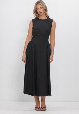 Frau mit blondem Haar, die ein ärmelloses schwarzes Midikleid und braune offene Schuhe trägt, steht vor einem schlichten weißen Hintergrund.