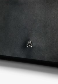 Bolso de cuero negro que presenta un emblema de una calavera y huesos cruzados en plata como acento distintivo, superficie texturizada y forma rectangular.