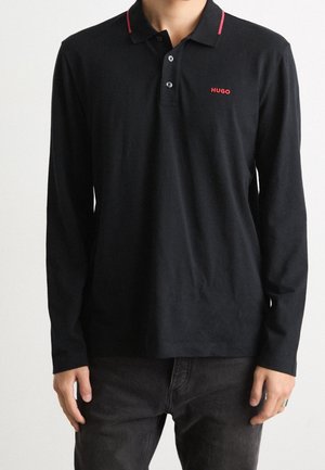 Person iført en sort langærmet polo med rød "HUGO"-logo på brystet og rødt kantbånd på kraven, kombineret med mørke bukser.