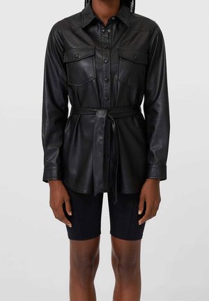 Femme portant une chemise noire en simili cuir à boutons avec poches poitrine et ceinture à la taille, associée à un short noir ajusté.