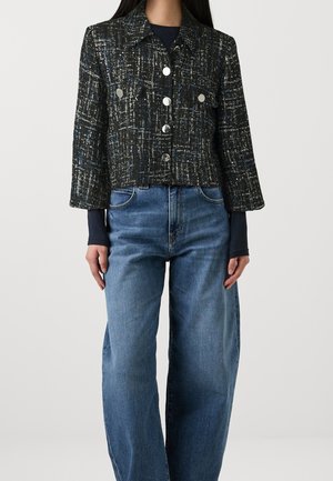 Femme portant une veste courte en tweed noir et blanc avec des boutons argentés, sur un haut à manches longues foncé, et un jean bleu décontracté.