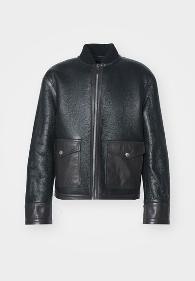 Zwarte leren bomberjack met een gestructureerde afwerking, geribbelde kraag, ritssluiting en twee voorzakken. Accenten in donkerbruin leer.