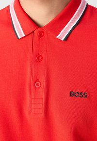 Polo rouge avec col rayé blanc et noir, trois boutons rouges, et logo "BOSS" noir brodé sur la poitrine.