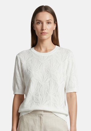 Donna con capelli castani lisci che indossa un maglione bianco a maniche corte con motivo di foglie e pantaloni beige chiaro su uno sfondo semplice.