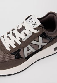 Primo piano di una sneaker marrone e grigia con lacci bianchi, suola texturizzata e grande logo A|X sul lato e sulla linguetta.