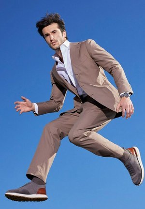 Abito color beige con una vestibilità aderente, camicia bianca e scarpe da ginnastica grigie con un accento marrone. Il modello è rappresentato in un salto contro uno sfondo blu.