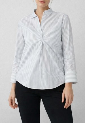 Donna che indossa una camicetta a maniche lunghe con righe verticali azzurre e bianche, con arricciatura sul davanti, e pantaloni neri.