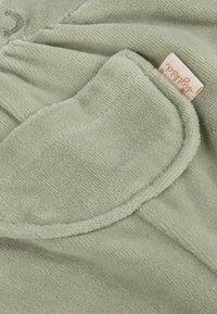 Soft, light green terry cloth fabric with a sewn-on rounded pocket and a beige fabric tag with cursive writing.