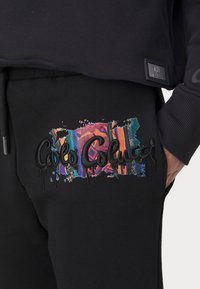 Svart genser med brodert fargerik logo som har sprut av lilla, blått og oransje, sammen med et strukturert stoffmerke.