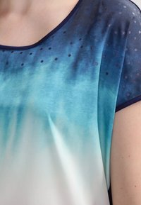 Blusa de manga corta con un degradado que va de azul oscuro a azul claro, con pequeños puntos azul marino a lo largo del escote, confeccionada en una tela ligera.
