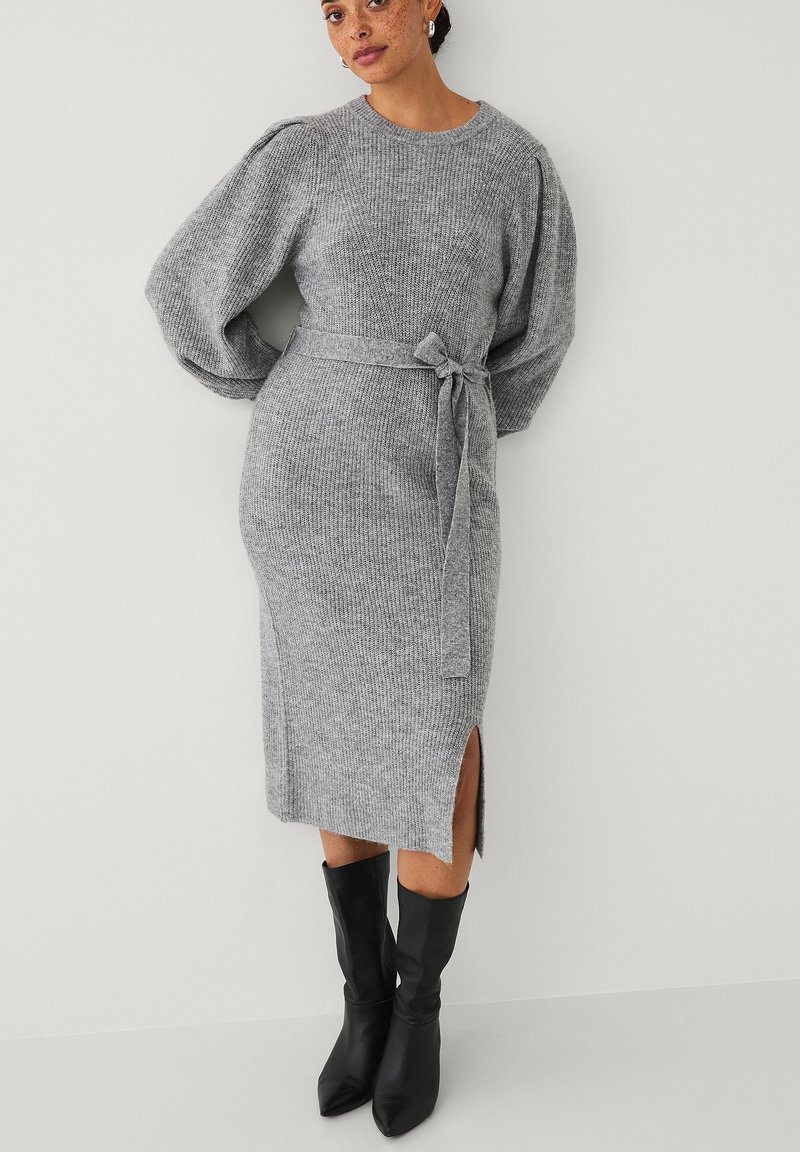Grijze gebreide midi-jurk met pofmouwen, een ceintuur in de taille en zijsplitten. Gepaard met zwarte laarzen metpuntige neuzen voor contrast.