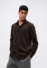 Brązowa koszula z długim rękawem, zapinana na guziki, z miękkiego materiału, z kołnierzykiem i listwą guzikową; noszona z szarymi, teksturowanymi spodniami.