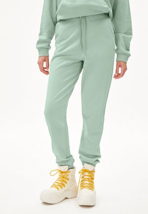Osoba ubrana w jasnozielone spodnie dresowe ze ściągaczem, w połączeniu z białymi wysokimi trampkami z żółtymi sznurowadłami, stojąca na białym tle.