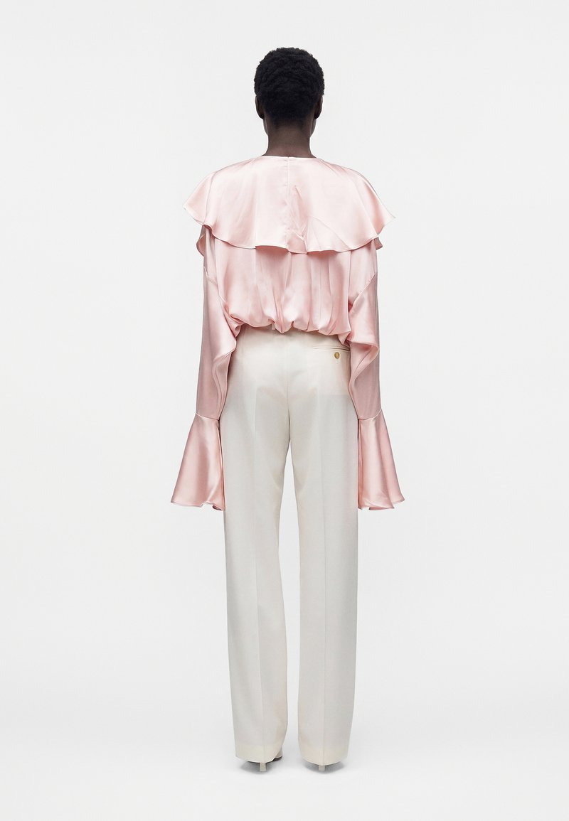 Personne portant une blouse satinée rose clair avec cape à volants et manches évasées, assortie à un pantalon blanc taille haute à jambes larges, debout.