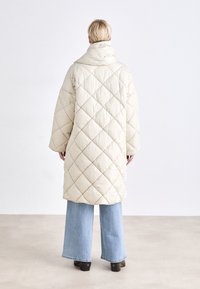 Casaco longo em creme, acolchoado, com gola alta, apresentando costura em forma de diamante e mangas largas, combinado com jeans azul claro de boca de sino.